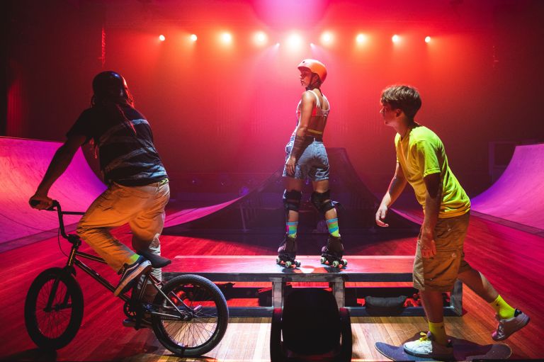 Three people on a stage with a skate ramp - one person on rollerskates, one on a BMX bike, and one on a skateboard